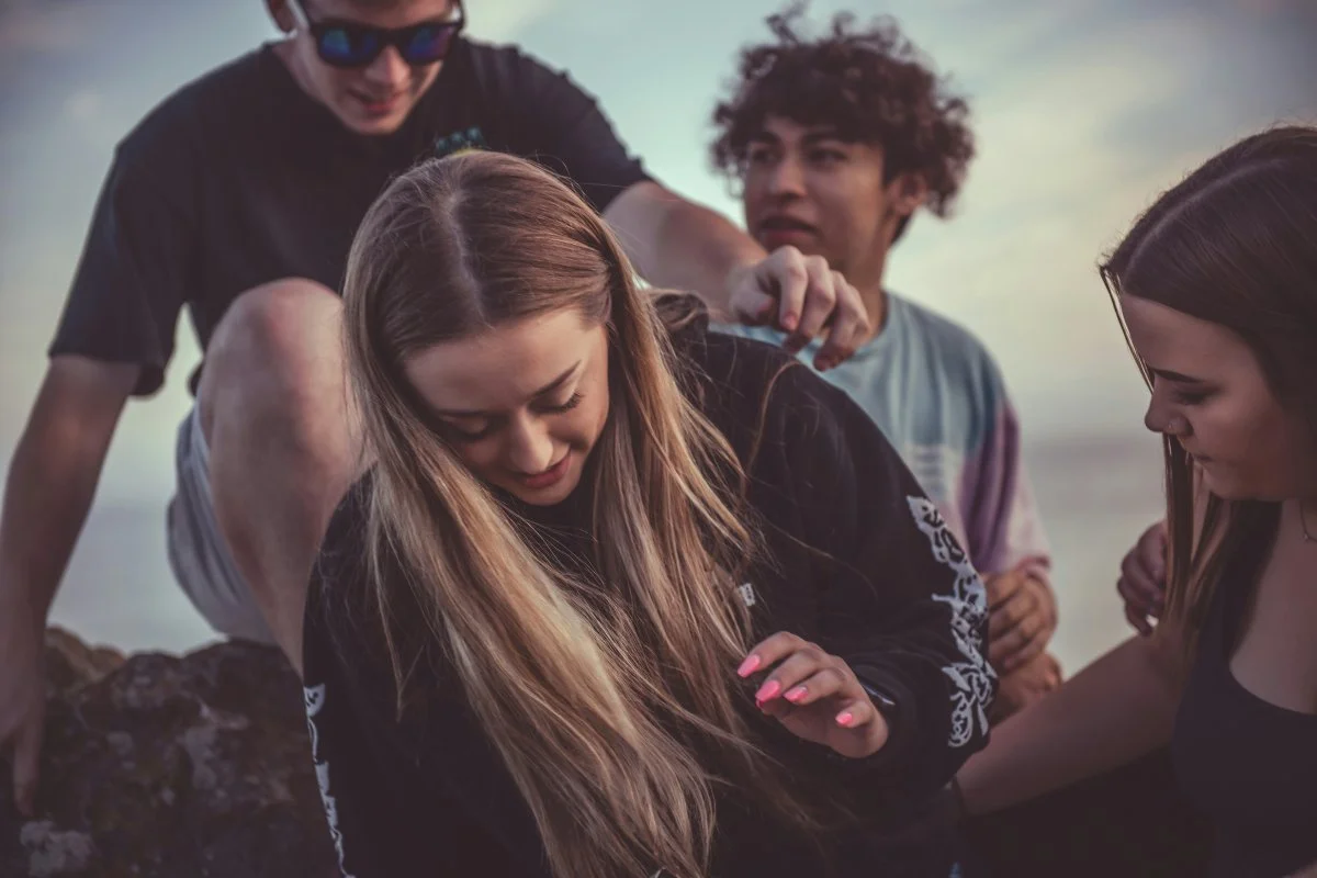 Groupe d'adolescents en quête d'apaisement Quatre adolescents discutant et s'installant sur des rochers en bord de mer, symbolisant le soutien, l'entraide et la quête d'apaisement face au refus scolaire.